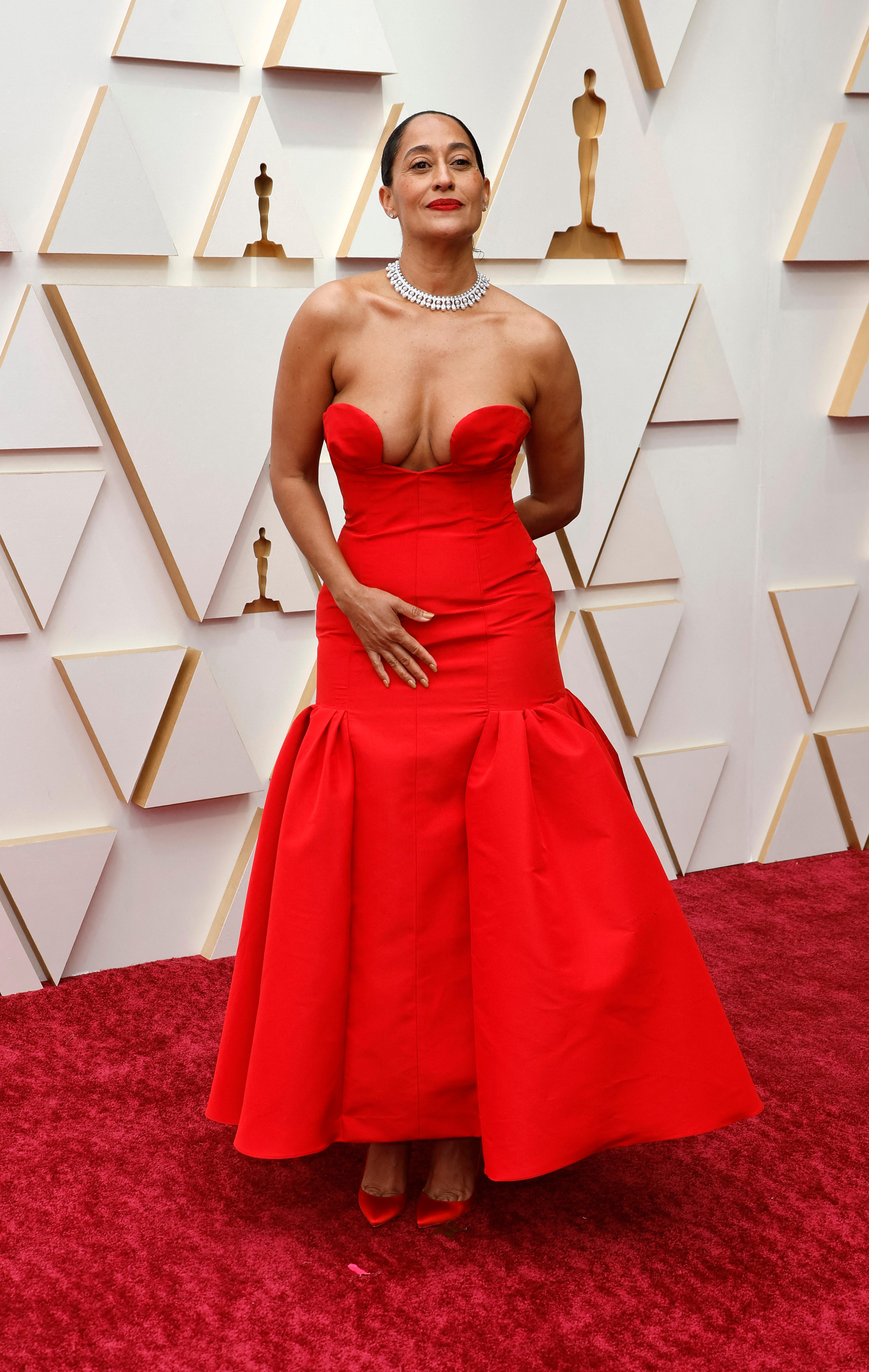 A woman wearing a long red dress stands on a red carpet in front of a white media wall for the Oscars.
