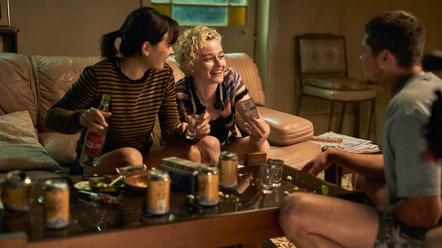 Two young women sit on a living room couch sharing a drink and a laugh with a young man sitting opposite.