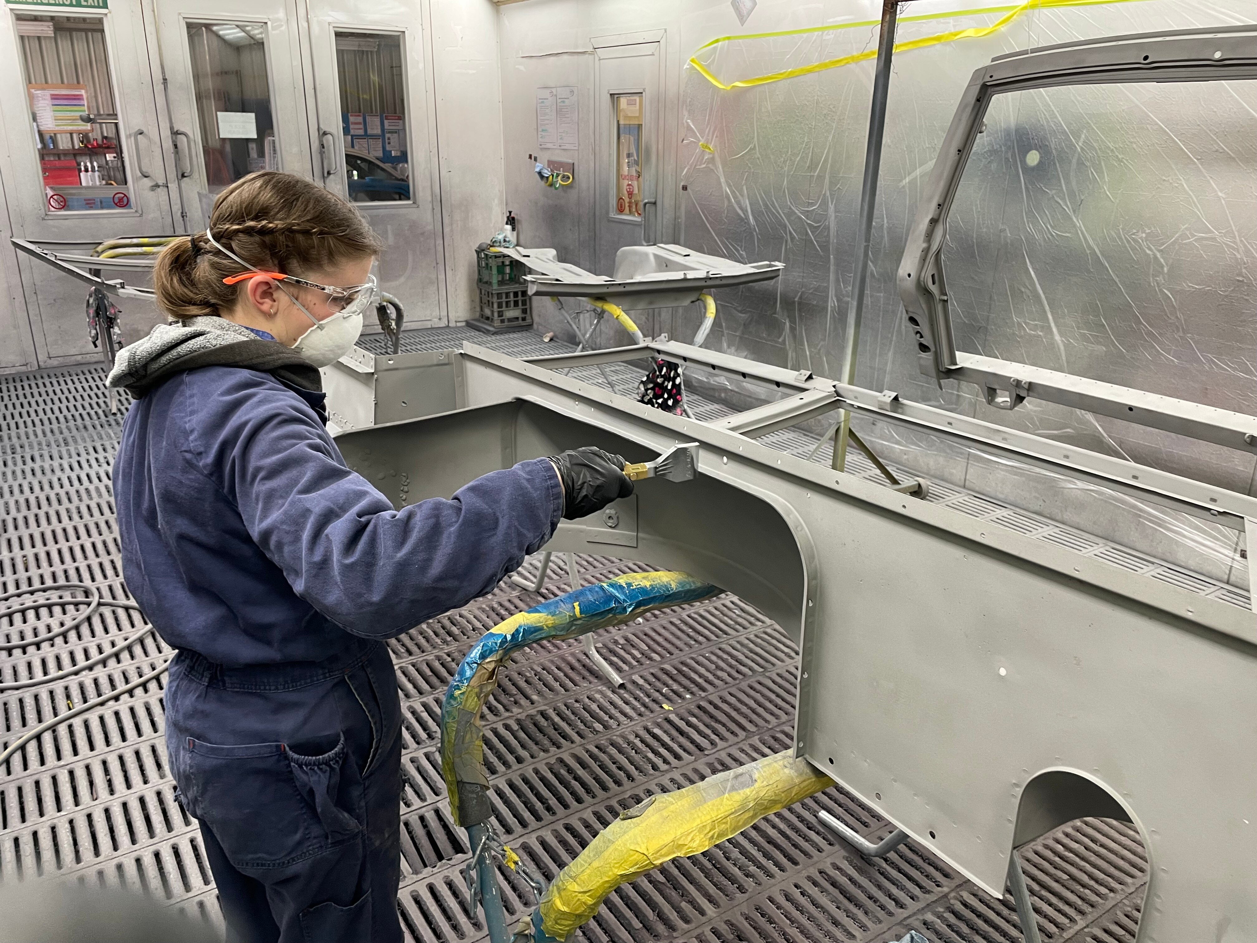 A girl in a blue overall, protective glasses, mask, uses a tool on a car part.