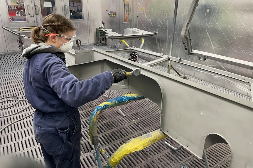 A girl in a blue overall, protective glasses, mask, uses a tool on a car part.