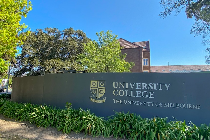 A black sign with the words "University College" and the "University of Melbourne" written on it in front of a large building.