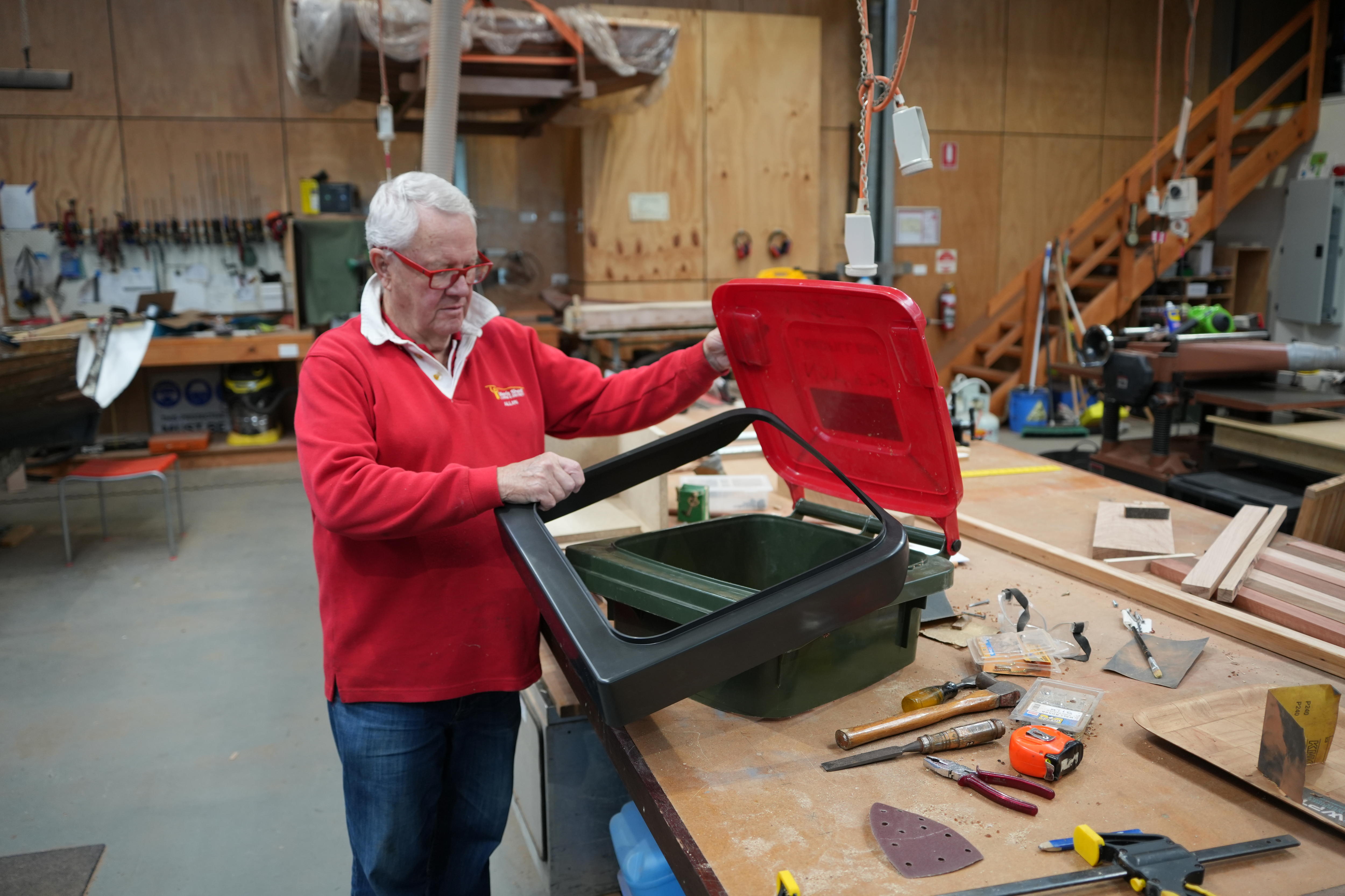 A white haired man in a red top and jeans holds a dark plastic square over the top of an open wheelie bin.