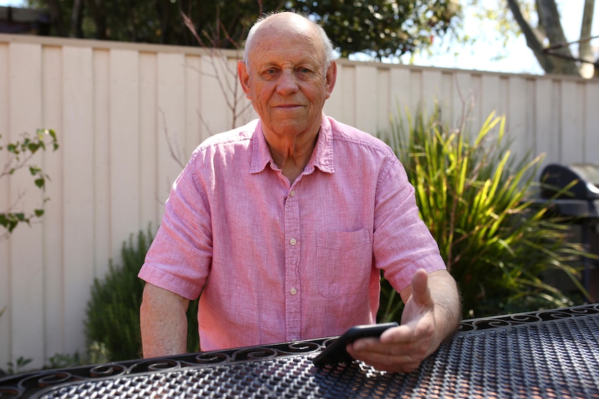 An older man in a pink shirt sits at an outdoor table.