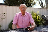 An older man in a pink shirt sits at an outdoor table.