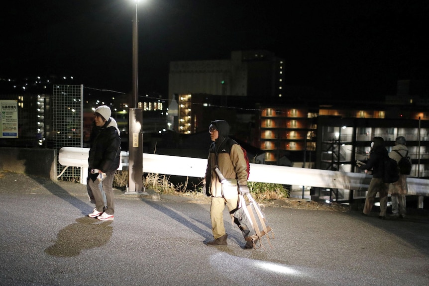 Two people in winter clothes carry bags up a paved road away from a town at night.
