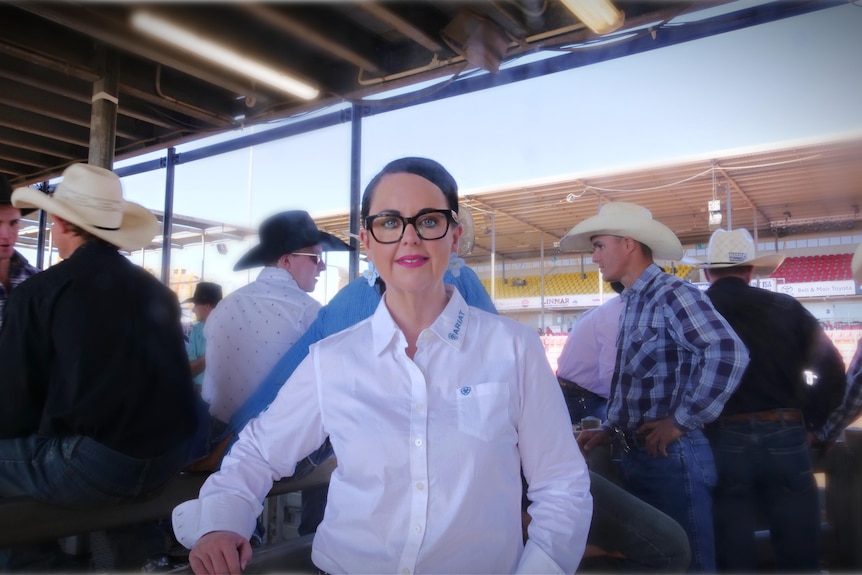 Mount Isa Rodeo chief executive Natalie Flecker stands in front of a group of cowboys. She wears a white rodeo shirt.