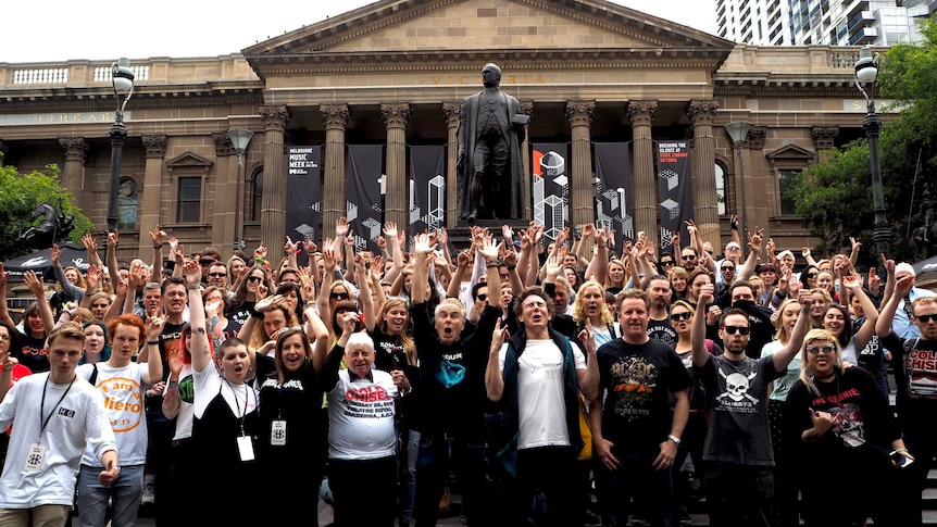 A large group of people wearing band t-shirts hold their arms in the air.