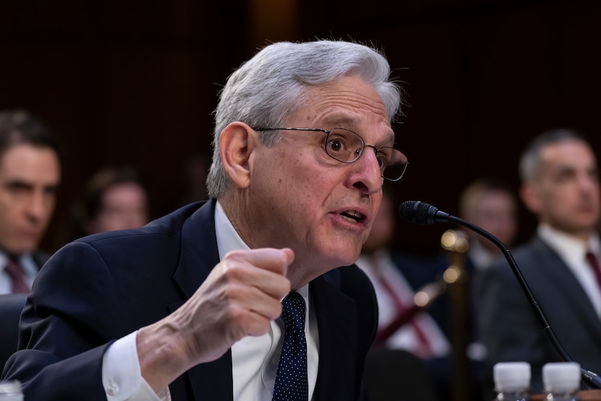 An older man with grey hair gestures as he speaks into a microphone in front of a seated crowd.