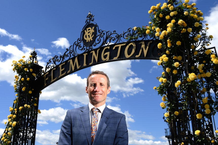 A suit-wearing jockey smiles as he stands under a racecourse entrance marked 'Flemington'.