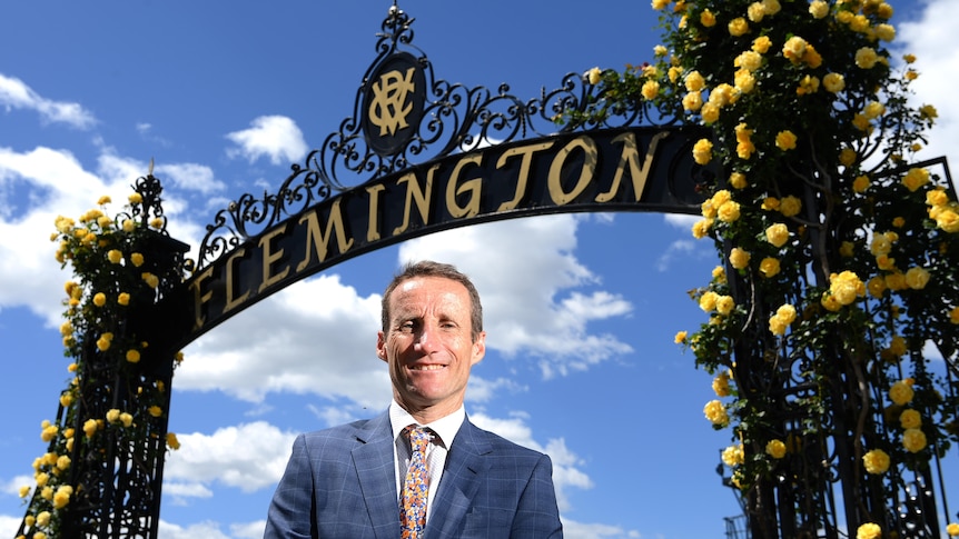 A suit-wearing jockey smiles as he stands under a racecourse entrance marked 'Flemington'.