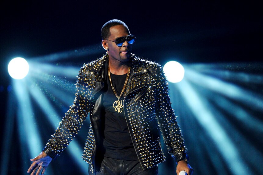 Man wearing sunglasses and dressed in black holds a microphone with bright lights shining behind him