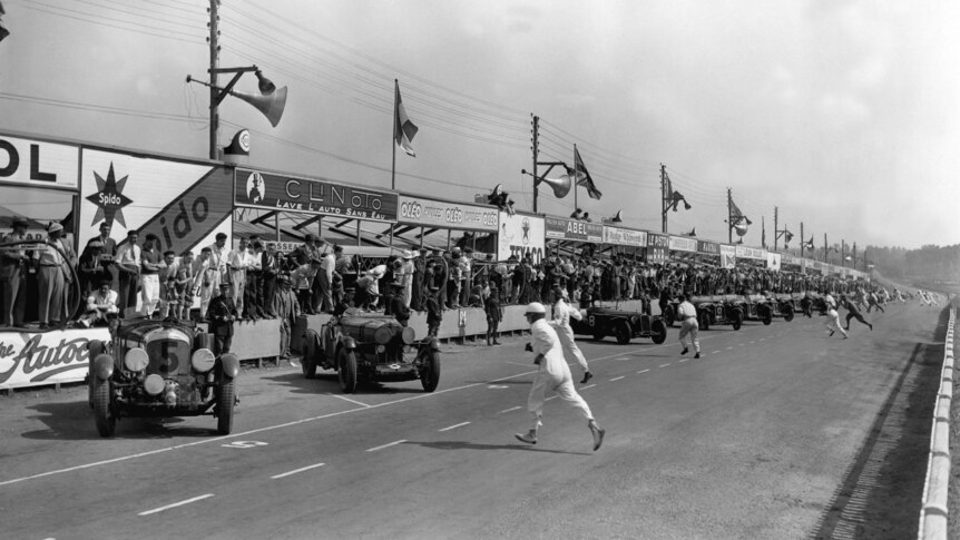 In this black and white photo, drivers rn to their cars to start a motor race. 