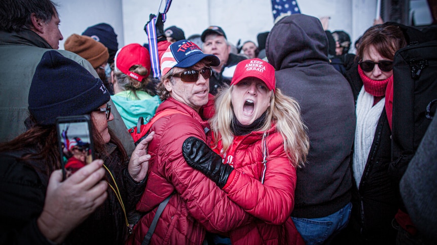 A woman in a Keep America Great hat shouts in a huge crowd of people holding US flags