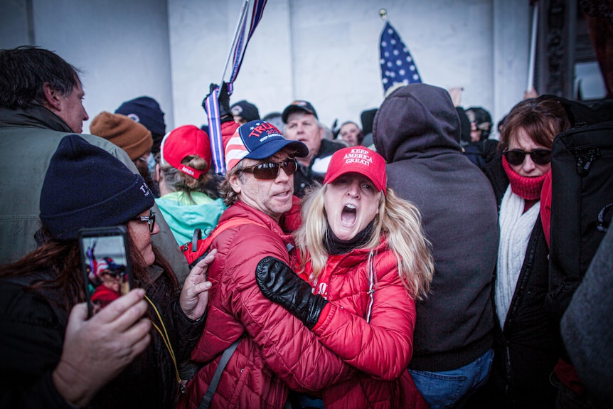 在一大群举着美国国旗的人群中，一名戴着 Keep America Great 帽子的妇女尖叫