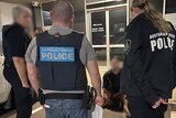 A man, face-blurred, sitting on a kerb out the front of a business surrounded by three AFP officers. 