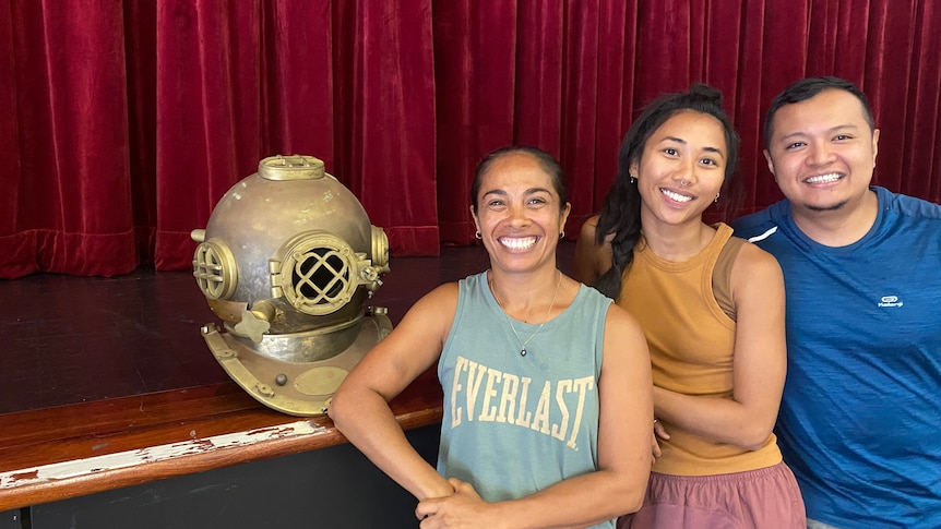 Two smiling women and a smiling man stand next to a replica pearl diving helmet in front of a stage