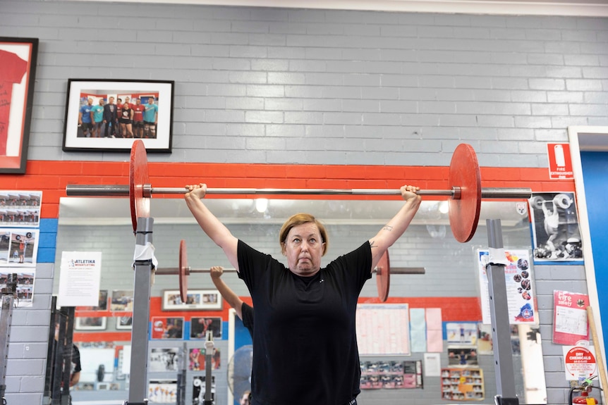 A middle-aged woman stands and lifts weights above her head.
