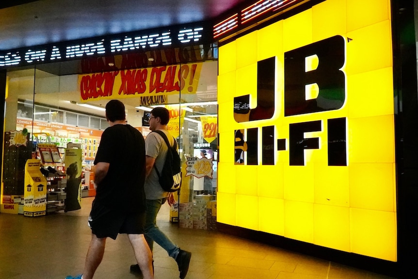 A JB Hi-FI sign outside a shop with two people walking past.