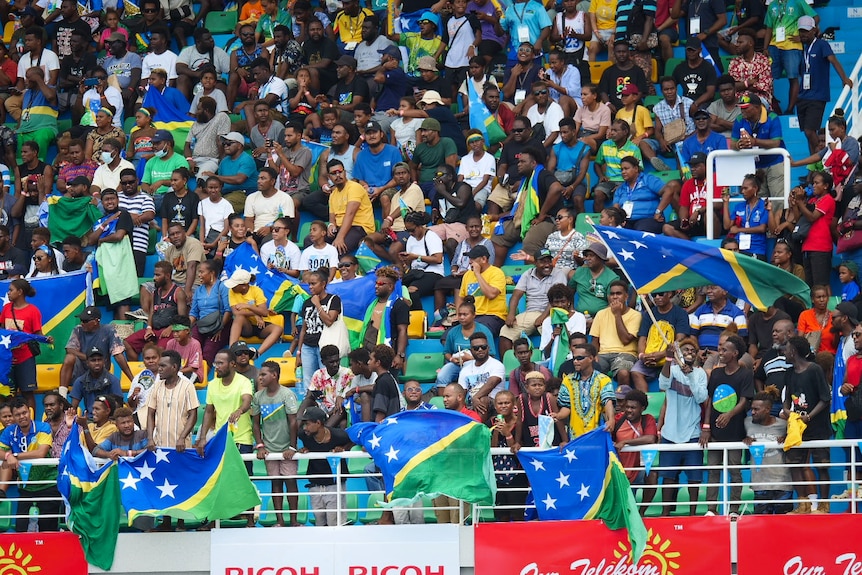 A crowd of people, many wearing Solomon Islands colours, pack a stand to watch the game.