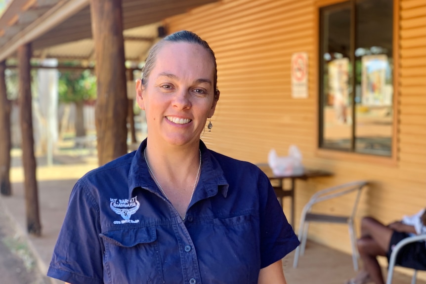 A woman in a blue shirt stands smiling in front of a verandah