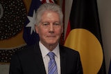 Smith, wearing a suit and standing in front of the Australian, Aboriginal and Torres Strait flags smiles tightly at the camera