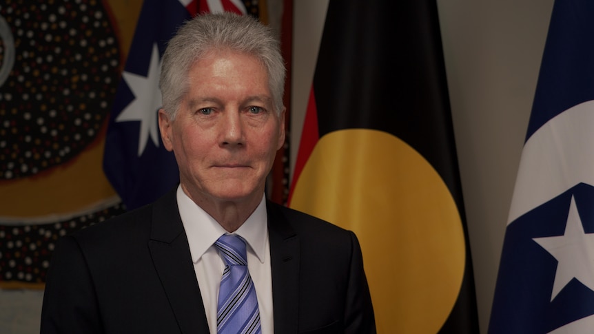 Smith, wearing a suit and standing in front of the Australian, Aboriginal and Torres Strait flags smiles tightly at the camera