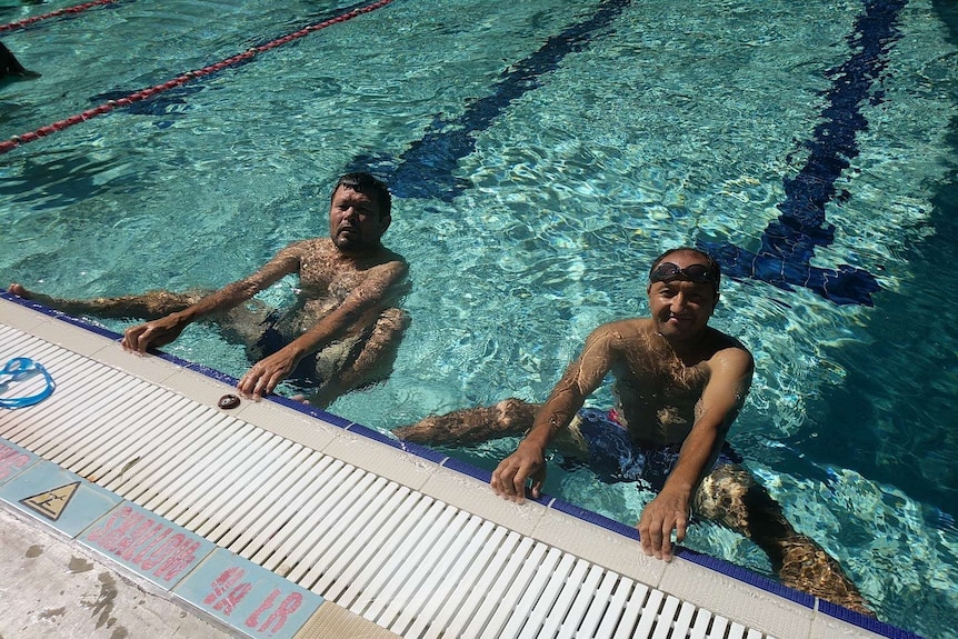two men in a pool holding on to the pool edge smiling