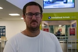 Blake Gilmore wearing glasses looks at the camera in front of the international arrivals section of Darwin Airport.