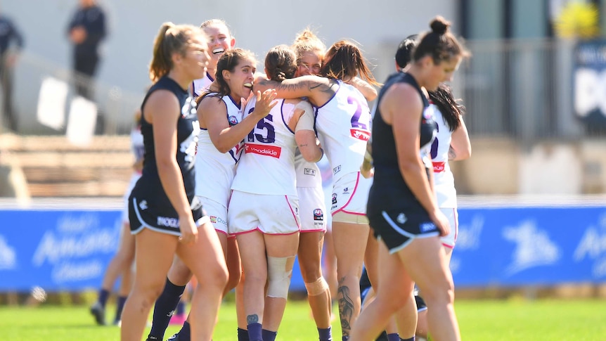 Fremantle Dockers hug after their AFLW win over Carlton, whose players are sad in the foreground.