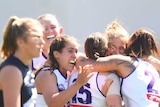 Fremantle Dockers hug after their AFLW win over Carlton, whose players are sad in the foreground.