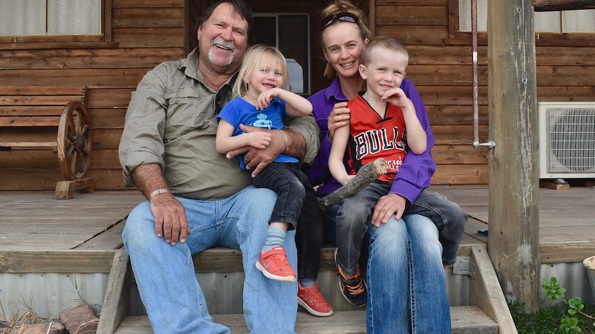 Family of four, two adults and two children, sit on the step of their house smiling and laughing with a wooden house in the back