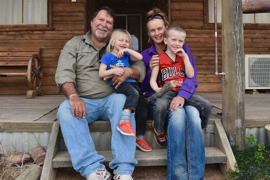 Family of four, two adults and two children, sit on the step of their house smiling and laughing with a wooden house in the back