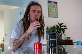 A man with dreadlocks, tattoos and a white shirt sits behind a table with cannabis paraphernalia, drawing on a vape.  