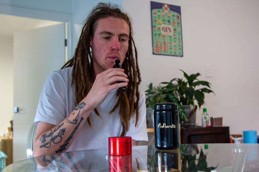 A man with dreadlocks, tattoos and a white shirt sits behind a table with cannabis paraphernalia, drawing on a vape.  