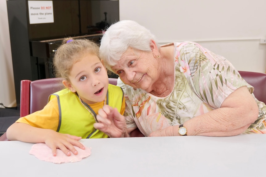 An elderly lady sits at a table leaning in near a young girl, smiling.