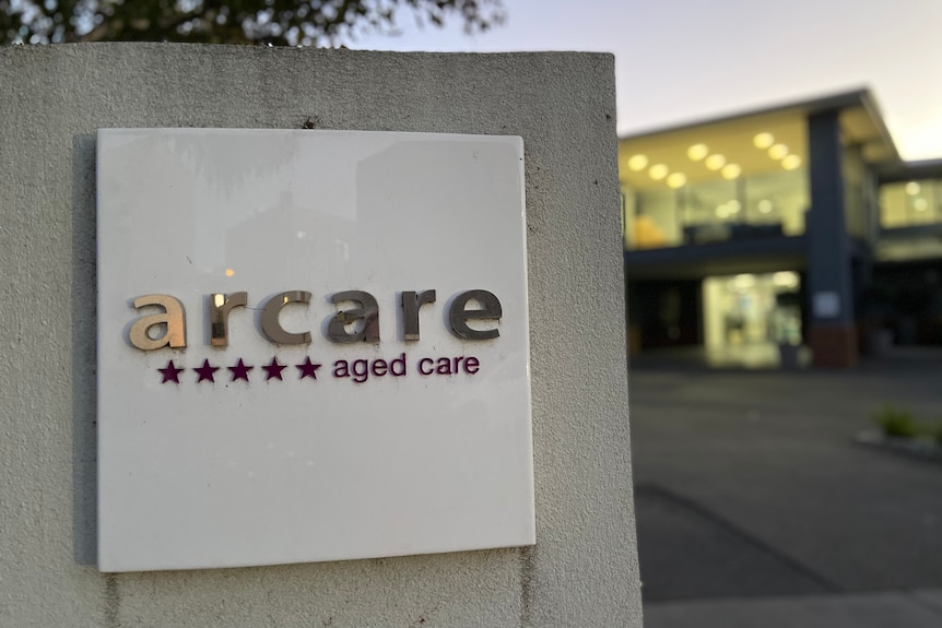 A sign in front of the home reads 'arcare aged care' with five stars, with lights on in the out-of-focus facility behind it.