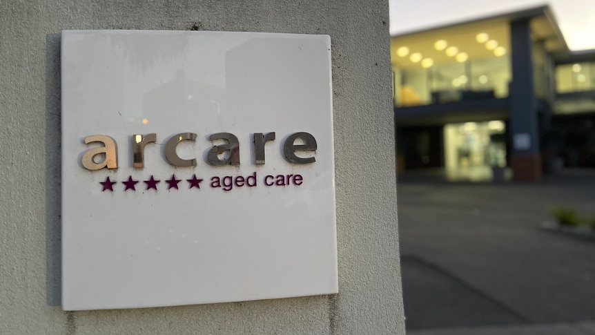 A sign in front of the home reads 'arcare aged care' with five stars, with lights on in the out-of-focus facility behind it.