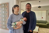 A woman holding a baby and her male partner stand in the doorway of their lounge room