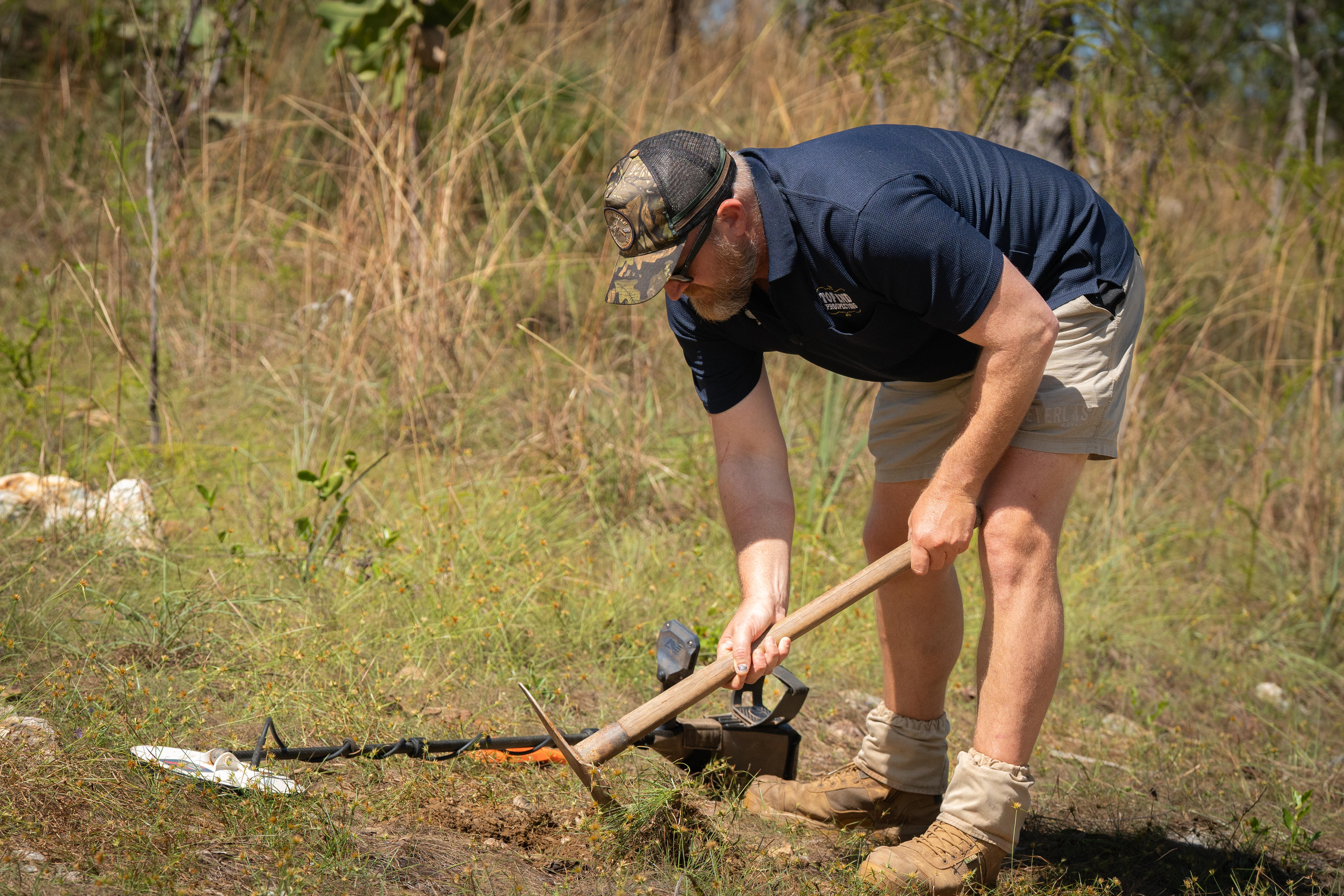 Man digs mattock into ground with detector next to him 
