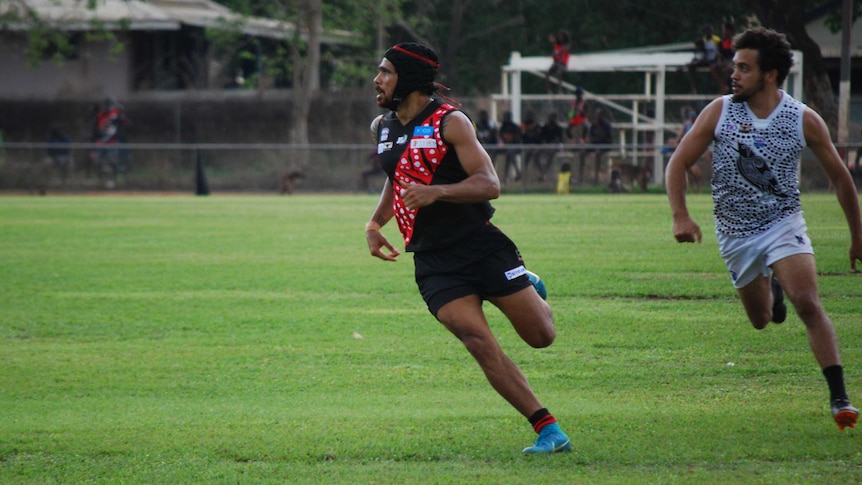 Cyril Rioli runs slightly in front of a Palmerston opponent on a grassy oval. Fans point in the background.