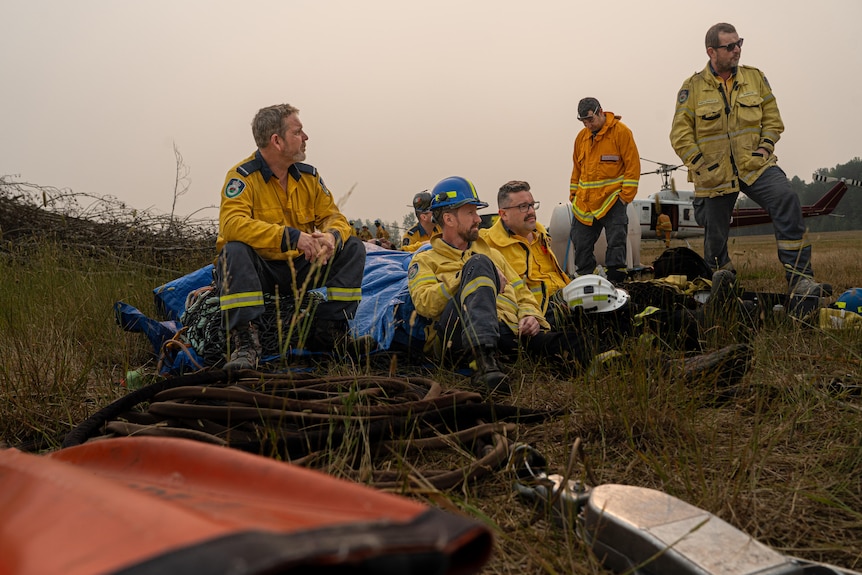 A group of firefighters in yellow uniforms sit around in a smoky field, waiting.