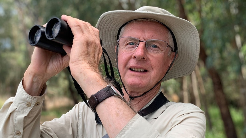 John Shepherd, at a wetlands, holds binoculars and looks beyond the camera lens.