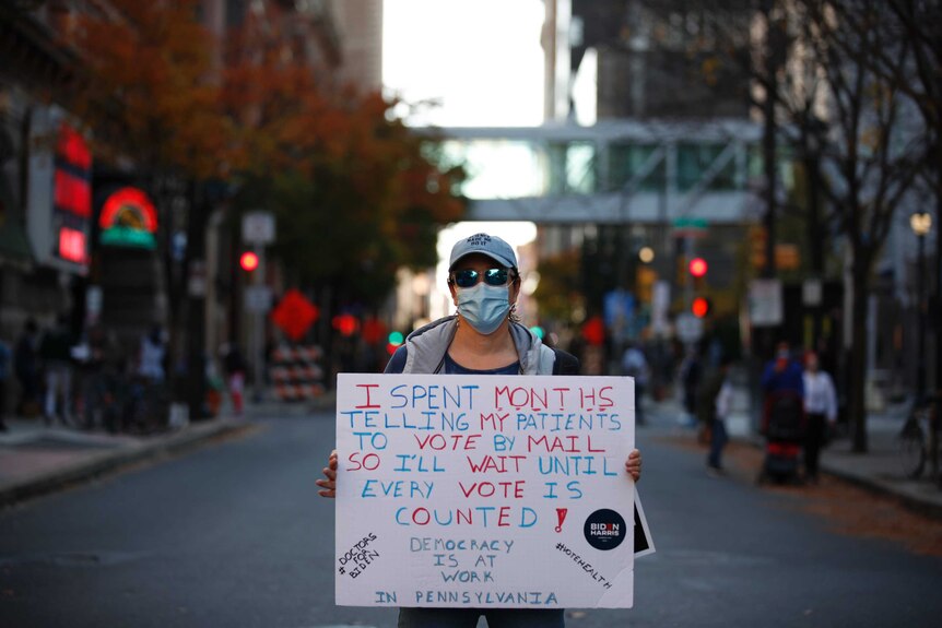 A doctor holds a placard referencing patients while standing outside a vote counting facility.