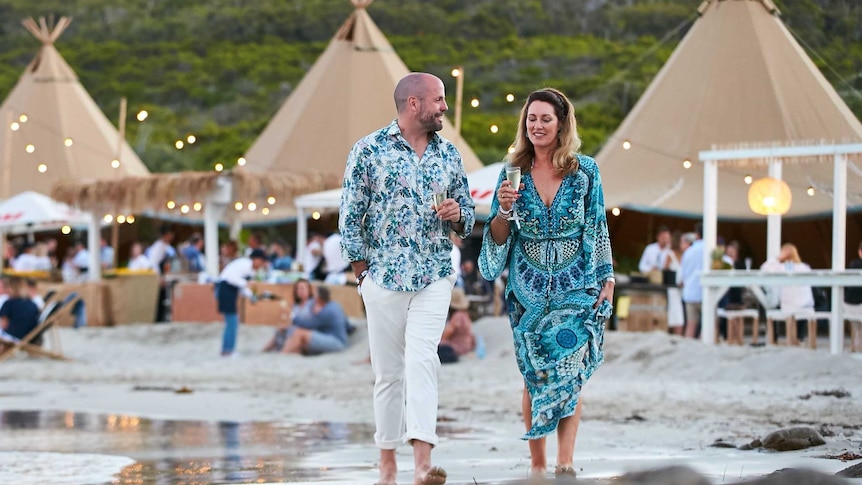 A man and woman are walking along the beach ion Margaret River, holding glasses of wine