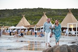 A man and woman are walking along the beach ion Margaret River, holding glasses of wine