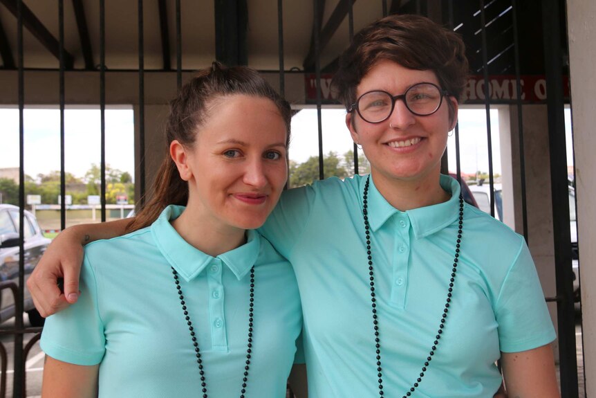 A mid shot of two young women wearing aqua polo shirts posing for a photo outside Fremantle Oval.