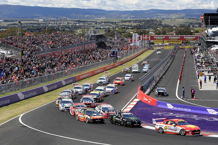 A number of cars approach and take a corner on a racetrack. A big crowd watches in the stands behind them.