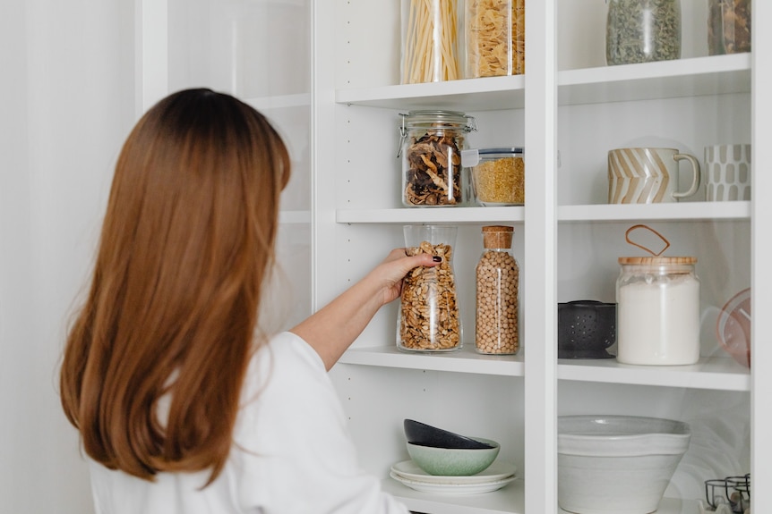 A woman with a white shirt and auburn hair organises glass jars of beans and pasta in her pantry