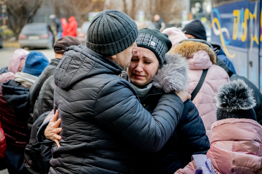 A man and woman in winter coats embrace. He hugs her close and kisses her cheek while she cries, she pulls him closer