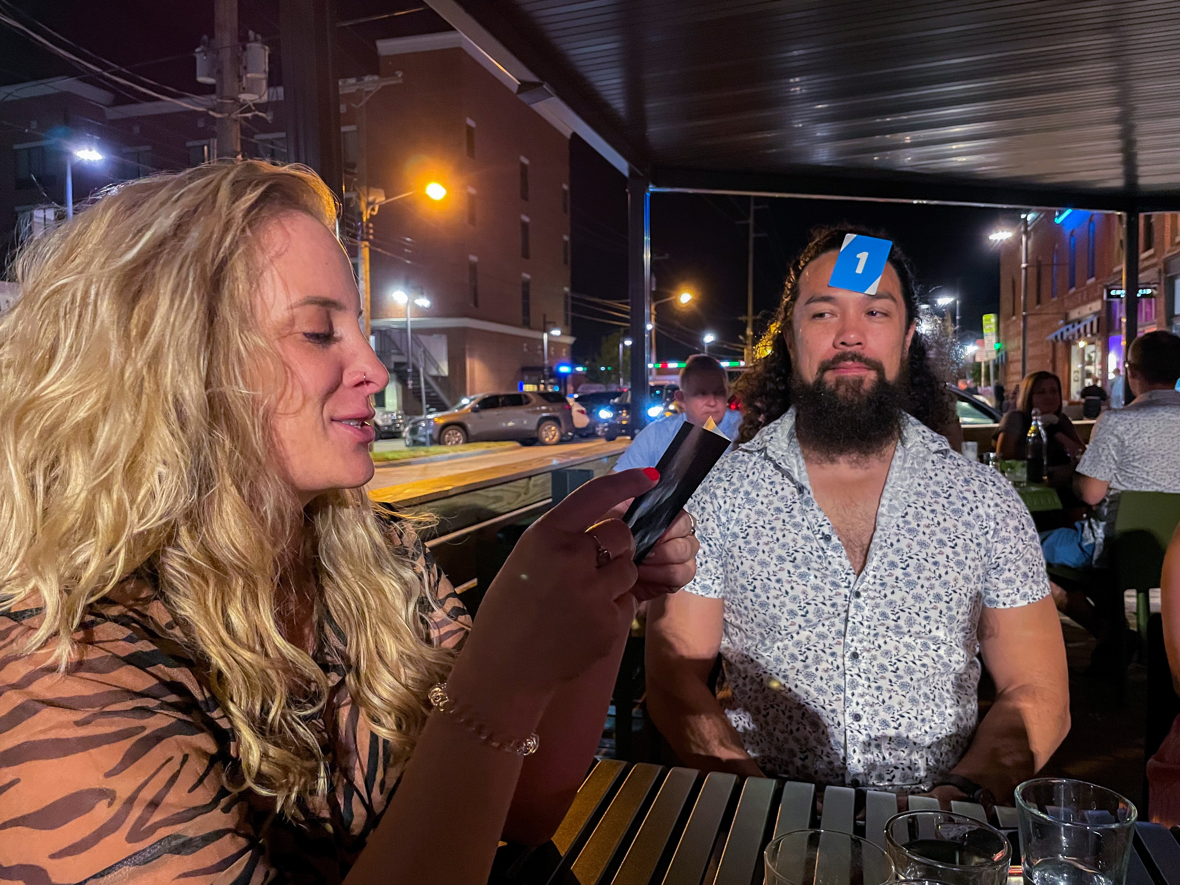 A man sits in a beer garden with a Uno card stuck to his forehead 